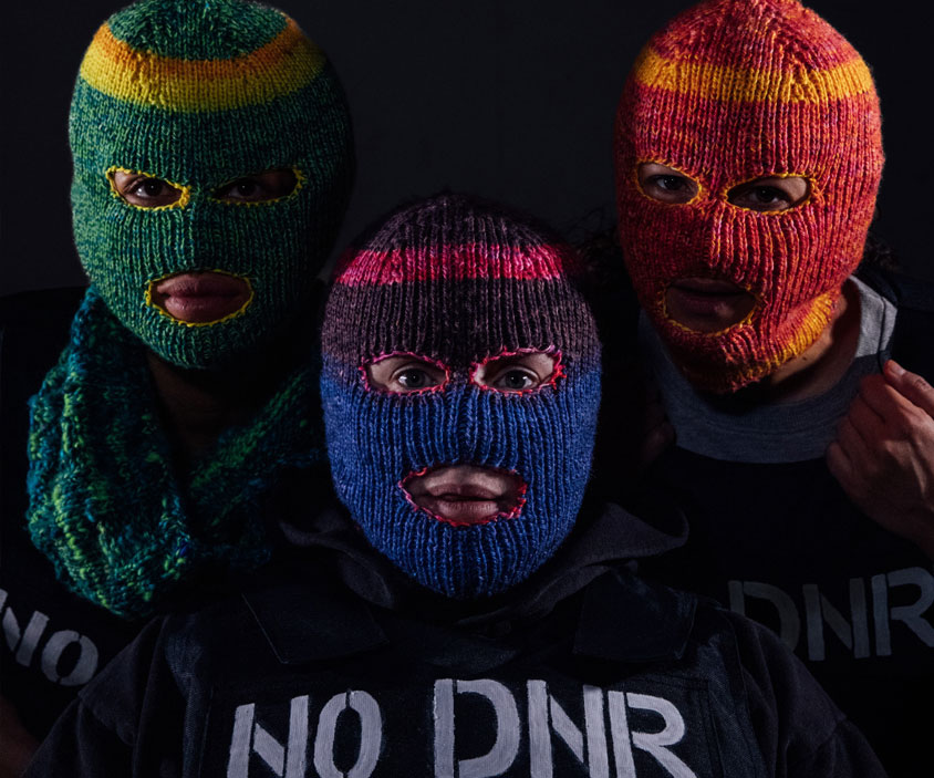 Three people stare directly at the camera and are each wearing different coloured, knitted balaclavas where only their eyes and mouths are exposed. They are all wearing black stab vests with the words ‘NO DNR’ stencilled onto the vests in white paint. Charmaine is on the left of the image. She is wearing a knitted green balaclava and blue knitted snood. Chopin is on the right of the image. She is wearing a red and orange knitted balaclava. She is tugging the top of the stab vest with her left hand. Jess is in the centre of the image between Charmaine and Chopin and is staring straight at the camera.  She is wearing a blue and pink knitted balaclava.